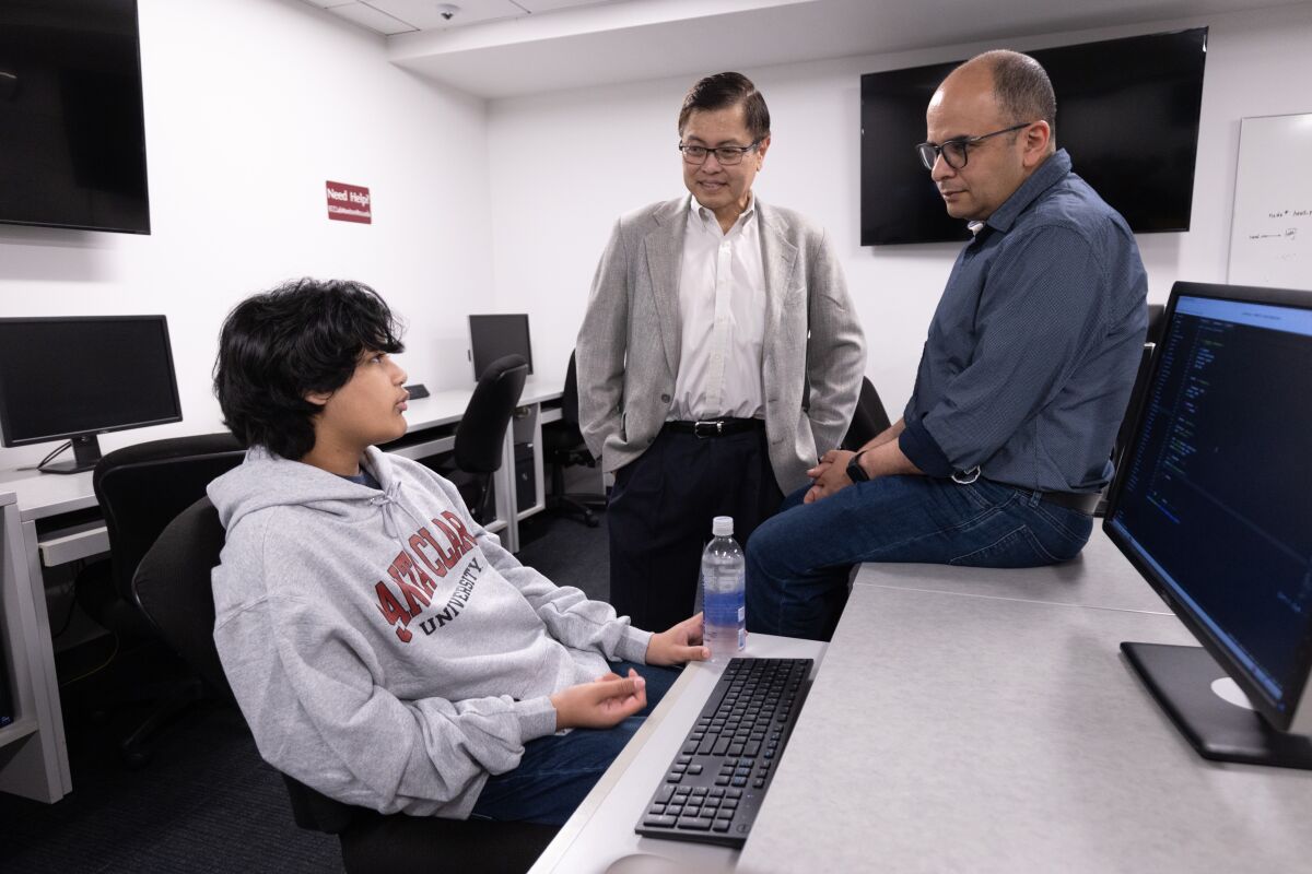 Кайран Квази, сидящий за столом с экраном компьютера, разговаривает с двумя мужчинами.